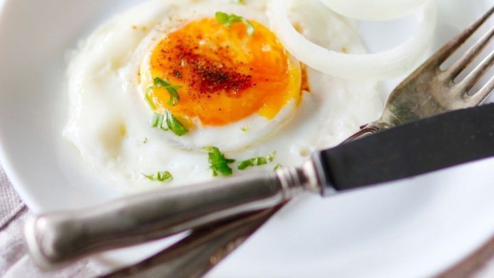 Ein perfekt gebratenes Spiegelei mit gelbem Eigelb liegt auf einem wei&szlig;en Teller, umgeben von Zwiebelringen und Kr&auml;utern.