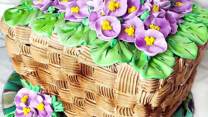 Ein kunstvoll dekorierter Kuchen mit lila Blumen und gr&uuml;nen Bl&auml;ttern steht auf einer Tortenplatte und sieht verf&uuml;hrerisch aus.