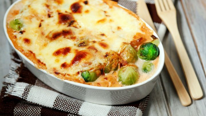 Ein Auflauf aus Rosenkohl ist goldbraun &uuml;berbacken und auf einem karierten K&uuml;chentuch platziert, umgeben von Besteck.