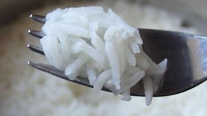 Ein L&ouml;ffel wei&szlig;er Reis wird auf einer Gabel gehalten, umgeben von dampfendem, lockerem Reis in einem Topf.