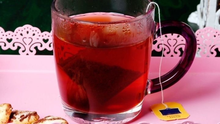 Ein Glas roter Tee mit Teebeutel steht auf einem rosa Tablett, umgeben von Pl&auml;tzchen, ideal zur Linderung bei Magen-Darm-Grippe.