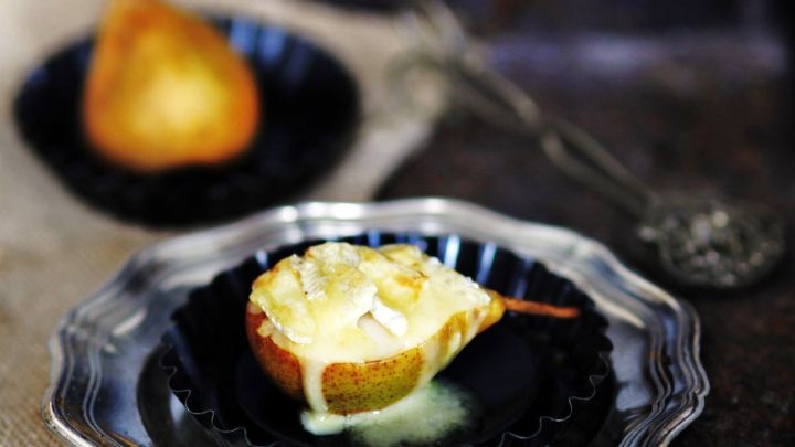 &Uuml;berbackene Birne mit Ziegencamembert auf einem silbernen Teller, umgeben von edlem Geschirr und einem rustikalen Hintergrund.