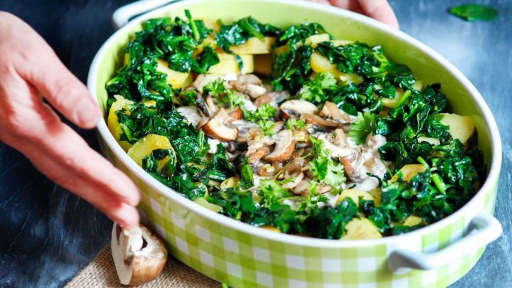 Ein gr&uuml;nes Gratin mit Spinat und Champignons wird von H&auml;nden gehalten, umgeben von frischen Zutaten auf einem Tisch.