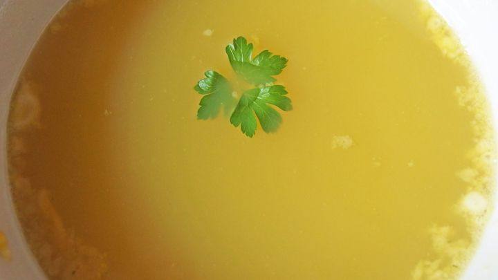 Eine goldene Einlaufsuppe mit einem frischen Kr&auml;uterblatt schwimmt in einer wei&szlig;en Sch&uuml;ssel.