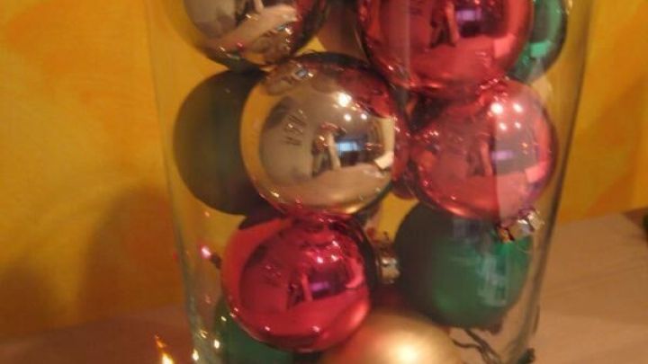 Christbaumkugeln in einer durchsichtigen Vase, umgeben von bunten Lichterketten, stehen auf einem Holztisch vor gelber Wand.