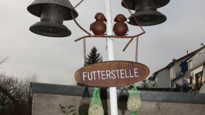 Ein Vogelhaus aus Resten h&auml;ngt als "FUTTERSTELLE" an einem wei&szlig;en Pfahl mit Glocken und zwei V&ouml;geln im Garten.