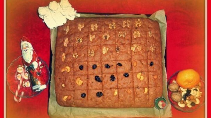 Honigkuchen mit N&uuml;ssen und Trockenfr&uuml;chten liegt auf Backpapier neben Weihnachtsdeko, Orange und N&uuml;ssen auf rotem Tuch.