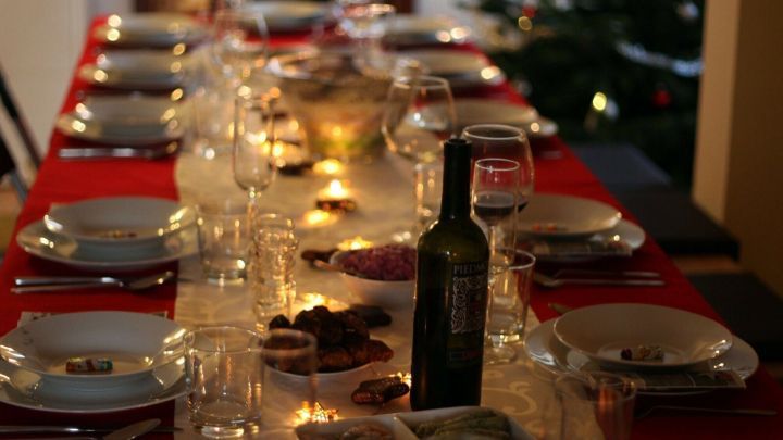 Ein festlich gedeckter Tisch mit Wein und Pl&auml;tzchen strahlt in einem gem&uuml;tlichen Raum, w&auml;hrend ein Weihnachtsbaum leuchtet.