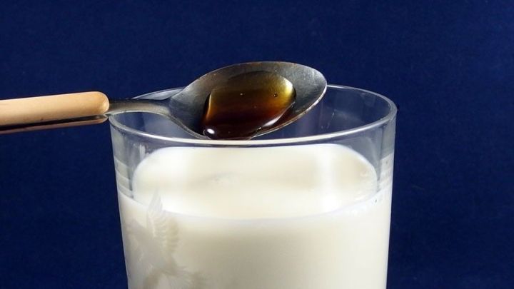 Ein L&ouml;ffel mit Honig schwebt &uuml;ber einem Glas Honigmilch, bereit zum Umr&uuml;hren, vor dunklem Hintergrund.