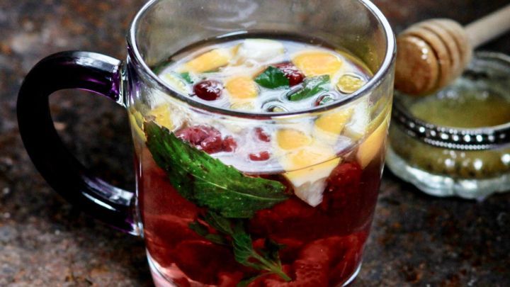 Ein durchsichtiger Becher mit frischem Fr&uuml;chtetee, gef&uuml;llt mit Fr&uuml;chten und Minze, steht neben einem Glas Honig.