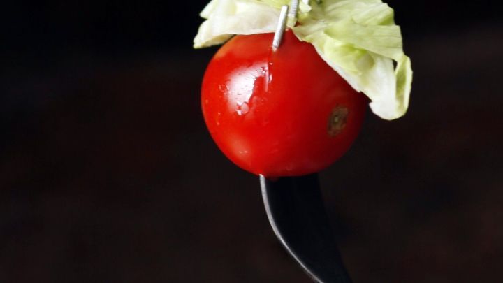 Eine Tomate mit einem gr&uuml;nen Blatt auf einer Gabel steht im Vordergrund, umgeben von einem unscharfen, dunklen Hintergrund.