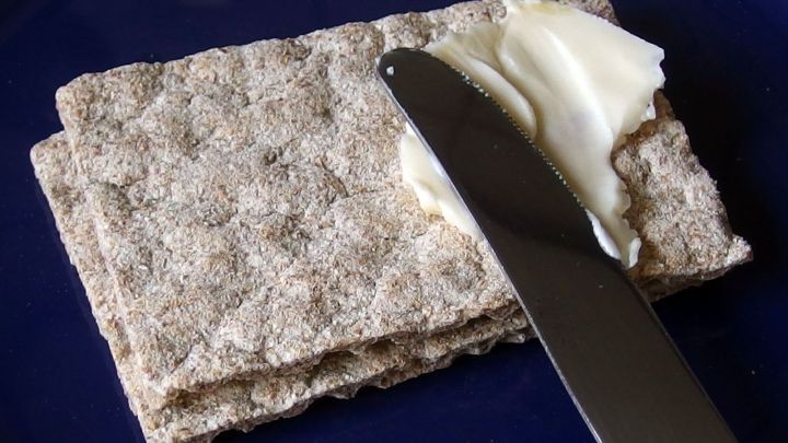 Kn&auml;ckebrot liegt auf einem blauen Teller, w&auml;hrend ein Messer darauf Butter verteilt, um es zu belegen.