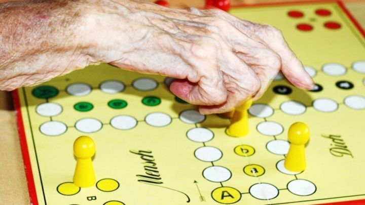 Eine alte Hand bewegt eine gelbe Spielfigur auf einem Brettspiel, um mit anderen Senioren Zeit zu verbringen.