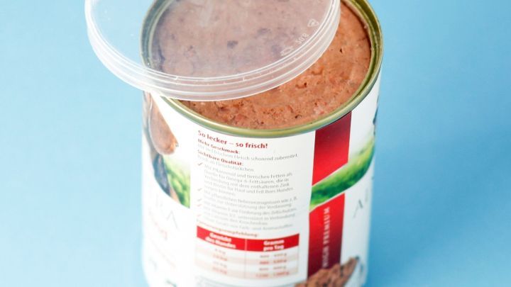 Eine halbge&ouml;ffnete Katzenfutter-Dose mit einem durchsichtigen Deckel auf blauer Unterlage zeigt frisches Futter.
