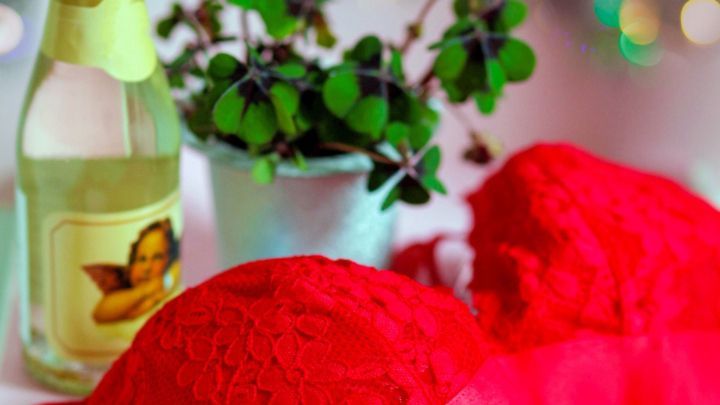 Rote Unterw&auml;sche liegt neben einer Flasche Sekt und einer Pflanze auf einem Tisch, dekoriert mit Lichtern im Hintergrund.