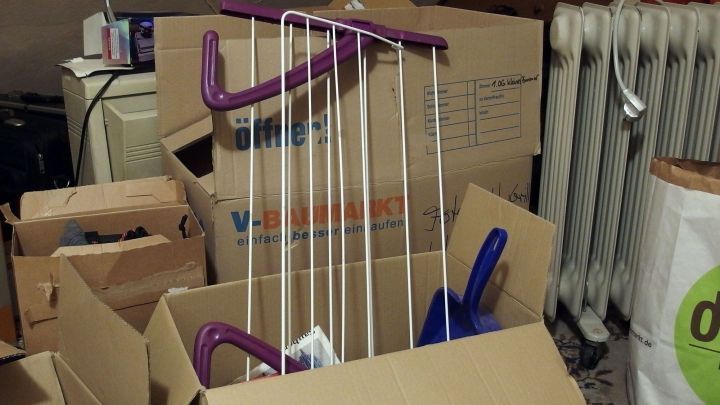 In einem &uuml;berf&uuml;llten Raum stehen mehrere Kartons, und ein W&auml;schegestell wird zum Trocknen von W&auml;sche genutzt.