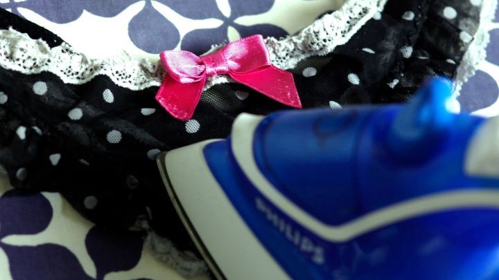 Ein blaues B&uuml;geleisen gl&auml;ttet den Kragen eines schwarzen, gepunkteten Kleides mit einem rosa Schleifenakzent.