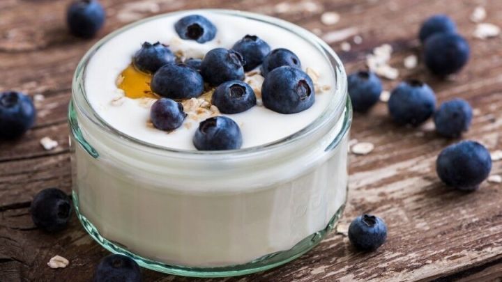 Ein Glas Sojajoghurt liegt auf einem Holztisch, garniert mit Heidelbeeren und etwas Honig, umgeben von weiteren Beeren.