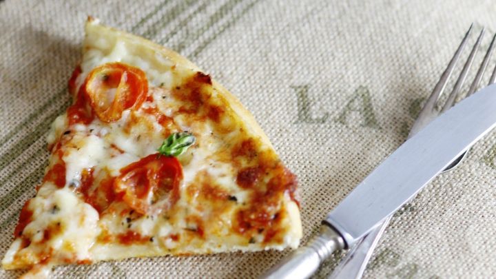 Eine Pizza mit saftigen Tomaten und K&auml;se liegt auf einem runden Gitterrost, w&auml;hrend ein St&uuml;ck vom Rand abgeschnitten ist.