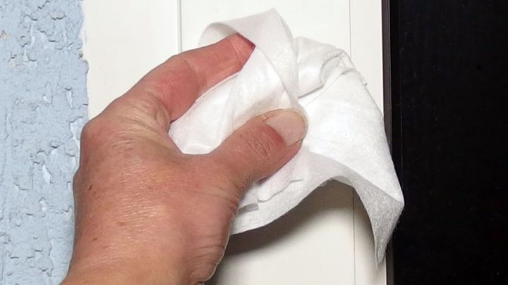 Eine Hand wischt mit einem Papiertuch an einem Fensterrahmen, um Nikotinflecken zu entfernen, vor einer Wand.