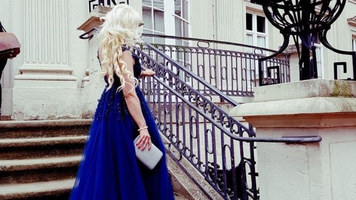 Eine Frau in einem langen, blauen Abendkleid geht elegant eine Treppe zu einem pr&auml;chtigen Geb&auml;ude hinauf.