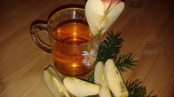 Eine Tasse mit Apfelpunsch steht auf einem Holztisch, umgeben von Apfelscheiben und Tannenzweigen.