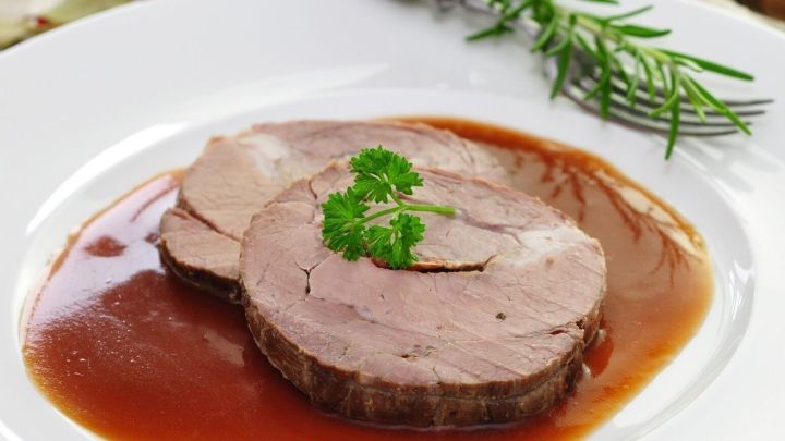 Zarte Fleischscheiben liegen auf einem Teller in reichhaltiger Bratenso&szlig;e, umgeben von Kr&auml;utern und einer rustikalen Tischumgebung.