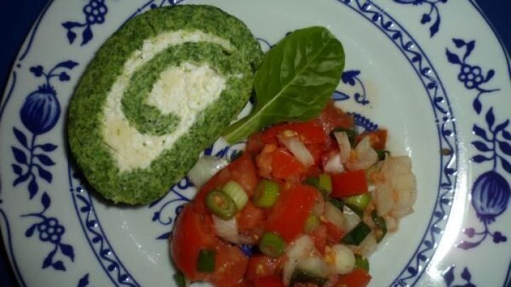 Eine gr&uuml;ne Biskuit-Spinatrolle liegt auf einem blauen Teller, umgeben von einem bunten Gem&uuml;sesalat und einem Blatt Spinat.