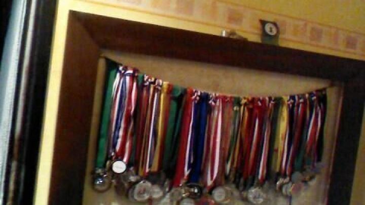 Ein Bilderrahmen h&auml;lt zahlreiche Medaillen mit bunten B&auml;ndern, aufgeh&auml;ngt an einer Wand in einem gem&uuml;tlichen Raum.