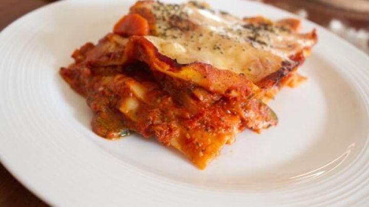 Eine appetitliche vegetarische Gem&uuml;selasagne liegt auf einem wei&szlig;en Teller, umgeben von einem rustikalen Tisch.