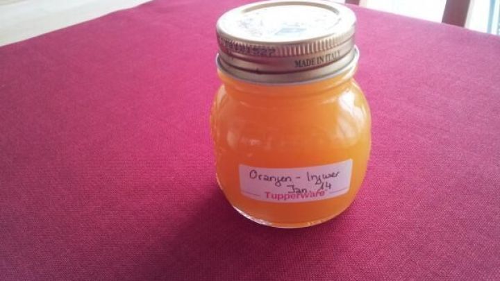 Ein Glas Orangen-Ingwer-Konfit&uuml;re steht auf einem roten Tisch. Das leuchtend orangefarbene Produkt ist in einem Schraubglas abgef&uuml;llt.