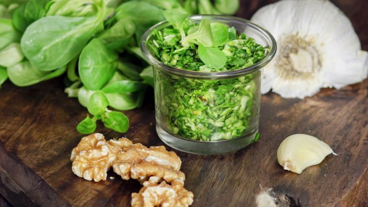 Feldsalat und Waln&uuml;sse liegen neben einem Glas mit gr&uuml;nem Pesto und einer Knoblauchzehe auf einem Holztisch.