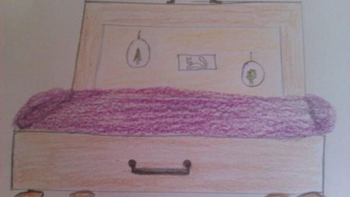 Ein Holzkoffer dient als Bett, ausgelegt mit einer lila Decke; an der R&uuml;ckseite h&auml;ngen Bilderrahmen mit B&auml;umen und einem Vogel.