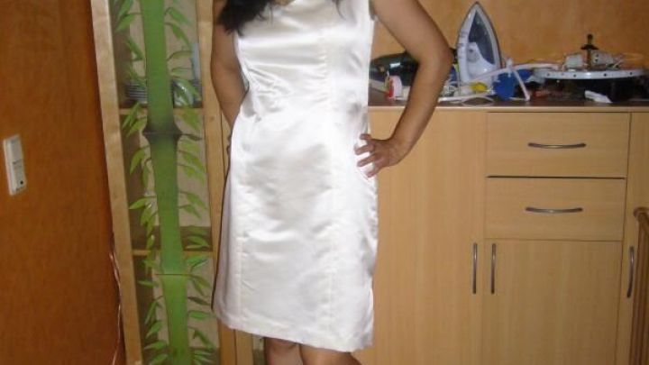 Eine Frau in einem eleganten wei&szlig;en Kleid posiert l&auml;chelnd in einem wohnlichen Raum mit Holzschrank und Wandkunst.