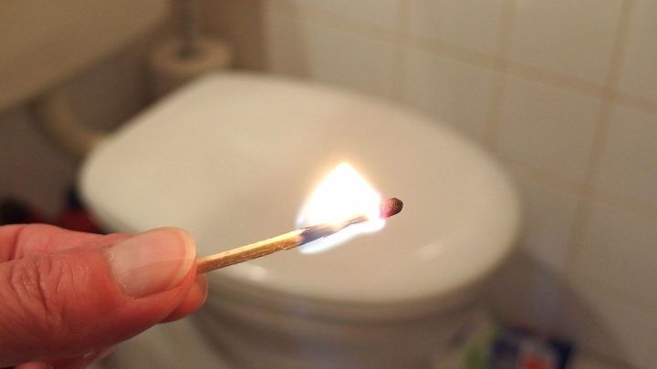 Eine brennende Z&uuml;ndholzstreichholz wird gehalten, w&auml;hrend es vor einer Toilette in einem Badezimmer entfacht wird, um Ger&uuml;che zu neutralisieren.
