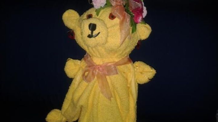 Ein gelber Handtuch-Teddy mit Blumenkranz und Schleife sitzt auf einem dunklen Sofa im Wohnzimmer.