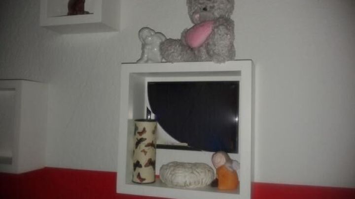 Ein Teddyb&auml;r sitzt auf einem Regal, umgeben von weiteren Spielzeugen, in einem Raum mit rot-wei&szlig;er Wandgestaltung.