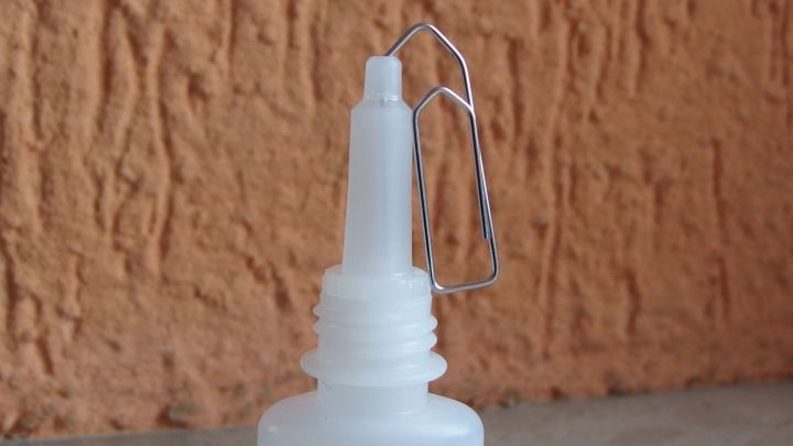 Eine Flasche Sekundenkleber steht vor einer strukturierten Wand, die T&uuml;lle wird durch einen Drahtverschluss freigehalten.