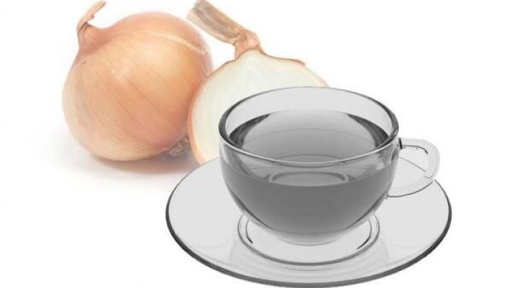 Eine Glastasse mit Zwiebeltee steht auf einem Unterteller, daneben liegt eine ganze und eine halbe Zwiebel auf wei&szlig;em Hintergrund.