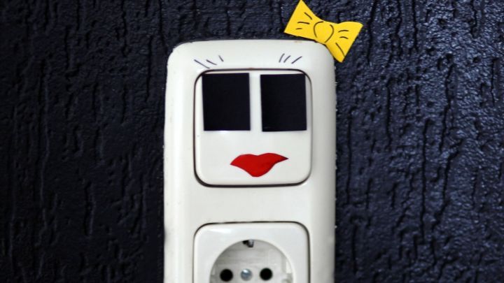 Ein dekorativer Lichtschalter in Form eines Gesichts mit roten Lippen und einem gelben Schleifchen, platziert an einer dunklen Wand.