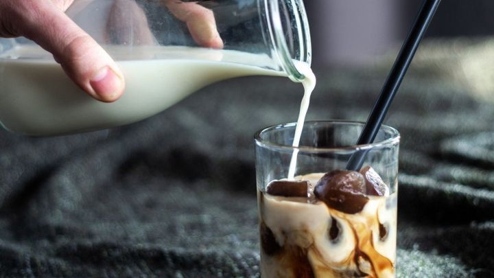 Eine Hand gie&szlig;t Milch in ein Glas mit Kaffee und Eisw&uuml;rfeln auf einem dunklen Tisch, was eine k&ouml;stliche Kaffeespezialit&auml;t ergibt.