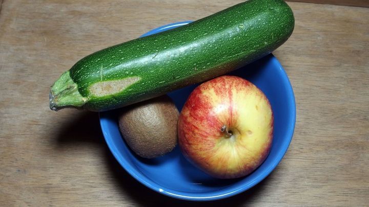 Eine gr&uuml;ne Zucchini, ein roter Apfel und ein Kiwi liegen in einer blauen Sch&uuml;ssel, umgeben von Holzoberfl&auml;che, reich an Vitaminen.