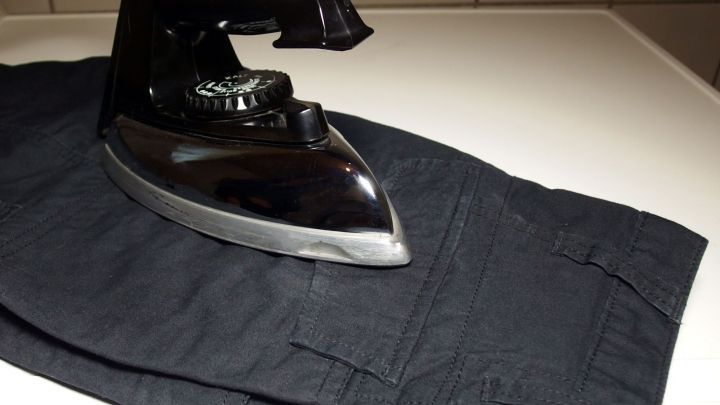 Ein schwarzer B&uuml;gel liegt auf einer schwarzen Hose, w&auml;hrend er im Hintergrund auf einer hellen Arbeitsfl&auml;che steht.