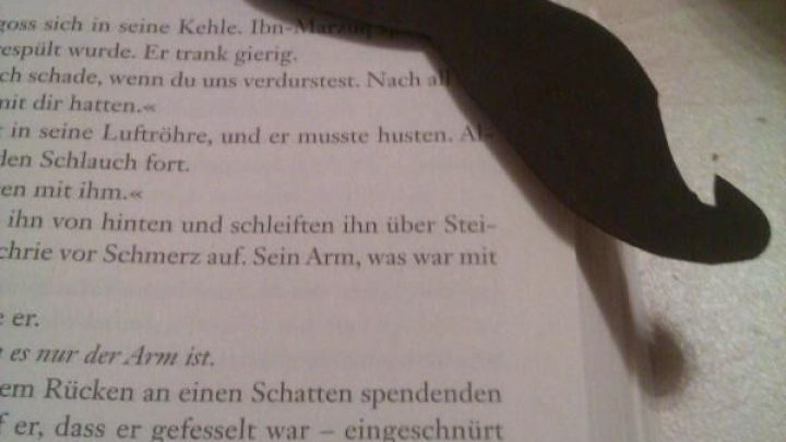 Ein schwarzes Mustache-Lesezeichen liegt auf einer offenen Buchseite, w&auml;hrend es eine Passage im Text markiert.