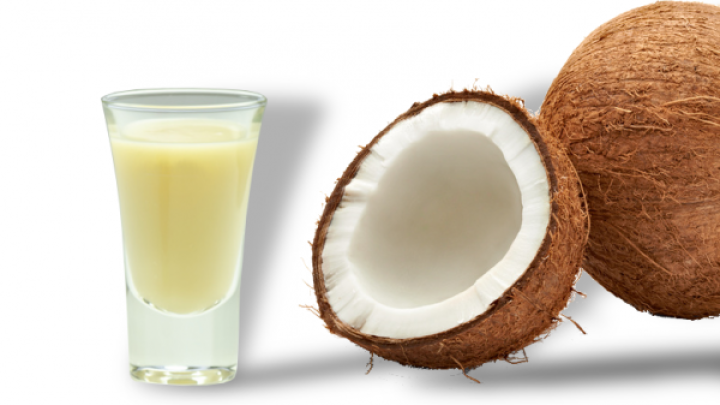 Ein Glas mit gelblicher Fl&uuml;ssigkeit steht neben zwei Kokosh&auml;lften auf wei&szlig;em Hintergrund.
