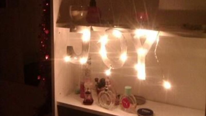 Beleuchtete Buchstaben "JOY" leuchten sanft auf einem Regal in einem dunklen Raum, umgeben von dekorativen Flaschen.