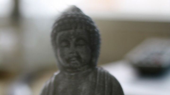 Eine Buddha-Statuette sitzt meditierend auf einem Tisch, umgeben von unscharfen Alltagsgegenst&auml;nden, die Ruhe symbolisieren.