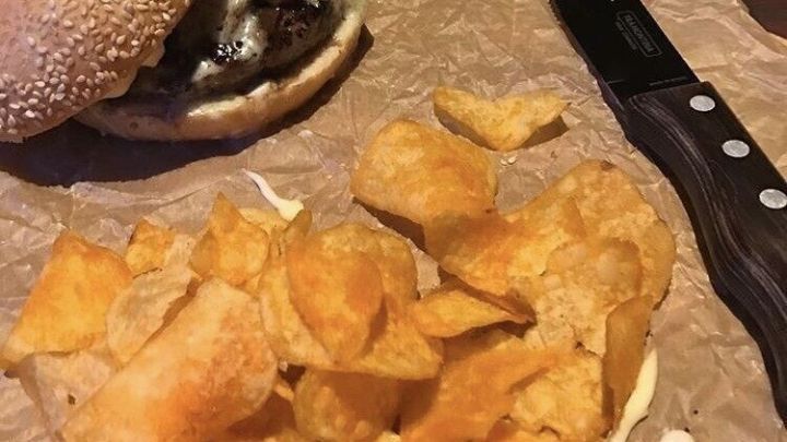 Ein Lamm-Burger liegt auf braunem Papier, umgeben von knusprigen Chips und einem Messer, bereit zum Genie&szlig;en.