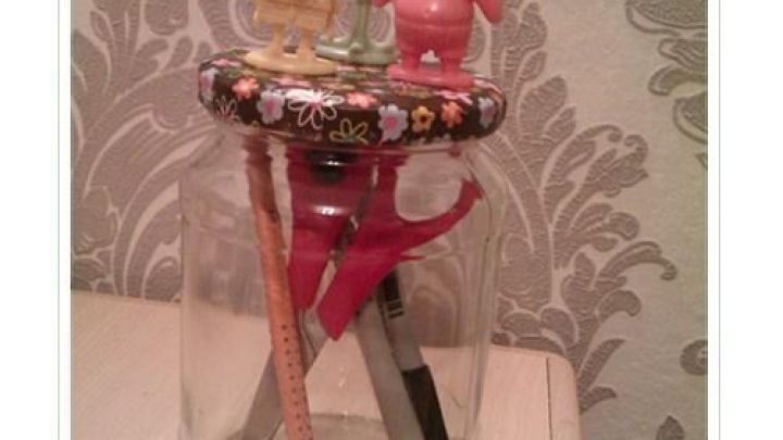 Ein Glas zur Aufbewahrung von Stiften mit dekorativem Deckel und Figuren, steht auf einem Tisch vor gemustertem Tapetenhintergrund.