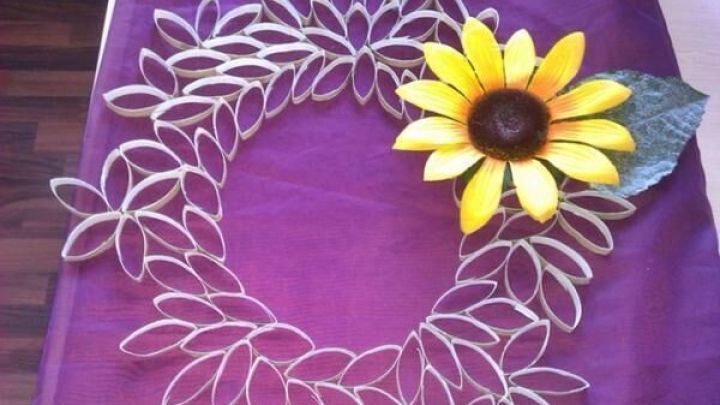 Ein dekorativer Kranz aus bl&uuml;tenf&ouml;rmigen Elementen liegt auf einem lila Untergrund, geschm&uuml;ckt mit einer Sonnenblume.